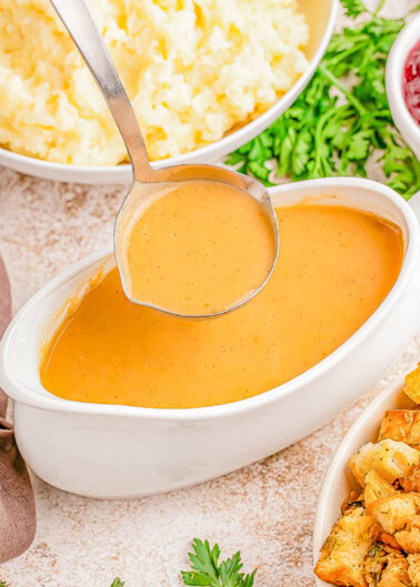 A ladle dips into a gravy boat filled with thick, brown gravy. Surrounding it are bowls of mashed potatoes and stuffing, garnished with parsley.