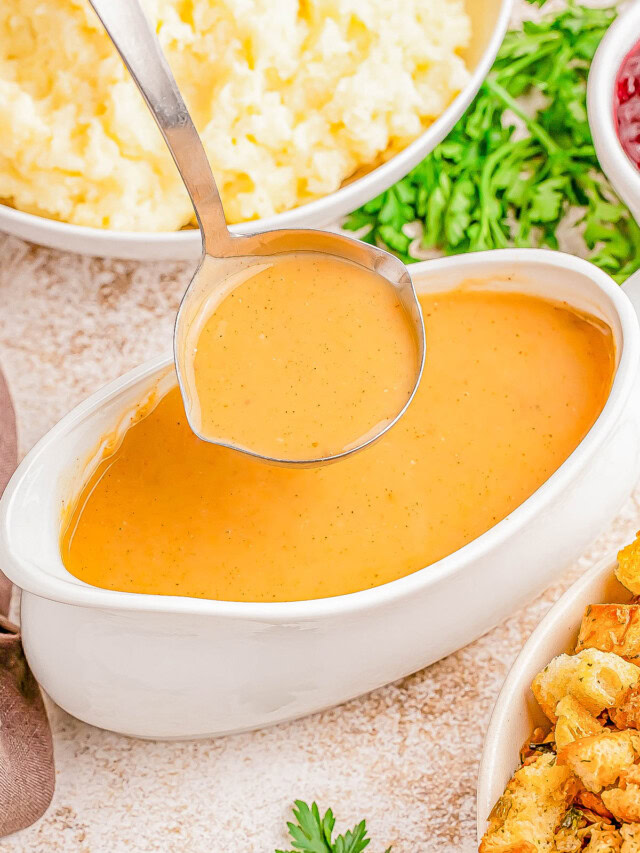 A ladle dips into a gravy boat filled with thick, brown gravy. Surrounding it are bowls of mashed potatoes and stuffing, garnished with parsley.