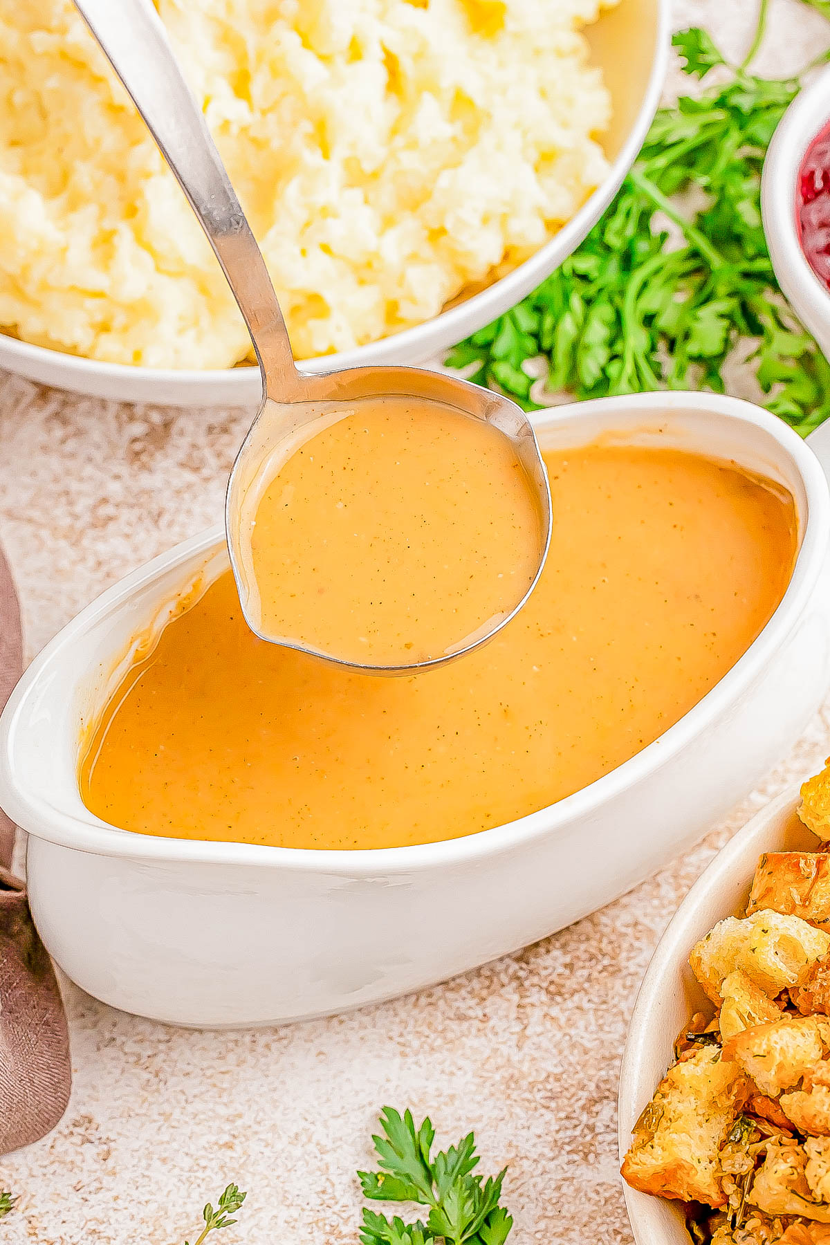 A ladle dips into a gravy boat filled with thick, brown gravy. Surrounding it are bowls of mashed potatoes and stuffing, garnished with parsley.