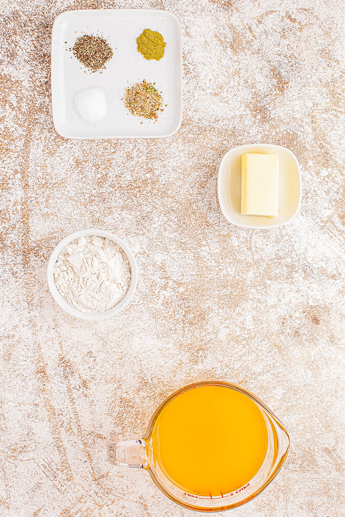 Ingredients on a countertop: small plates with spices, a bowl with butter, a bowl with flour, and a measuring cup with orange liquid (likely broth or juice).