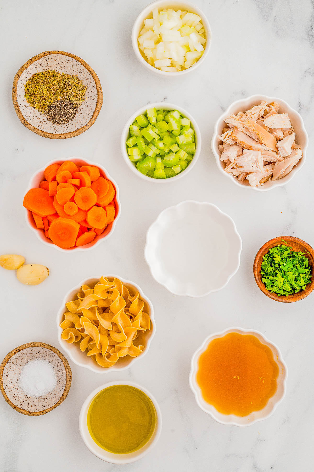 Ingredients for a soup arranged on a countertop include sliced carrots, chopped celery, diced onions, shredded chicken, egg noodles, broth, olive oil, salt, garlic, herbs, and parsley.