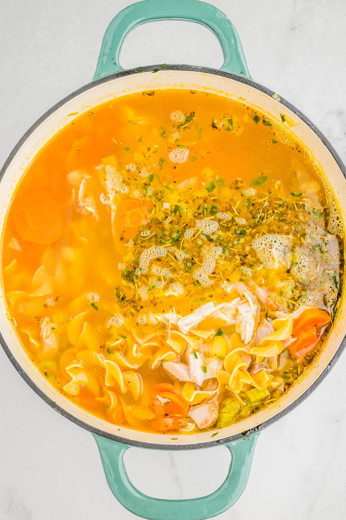 A pot of chicken noodle soup with carrots, herbs, and noodles in a broth, seen from above on a marble surface.