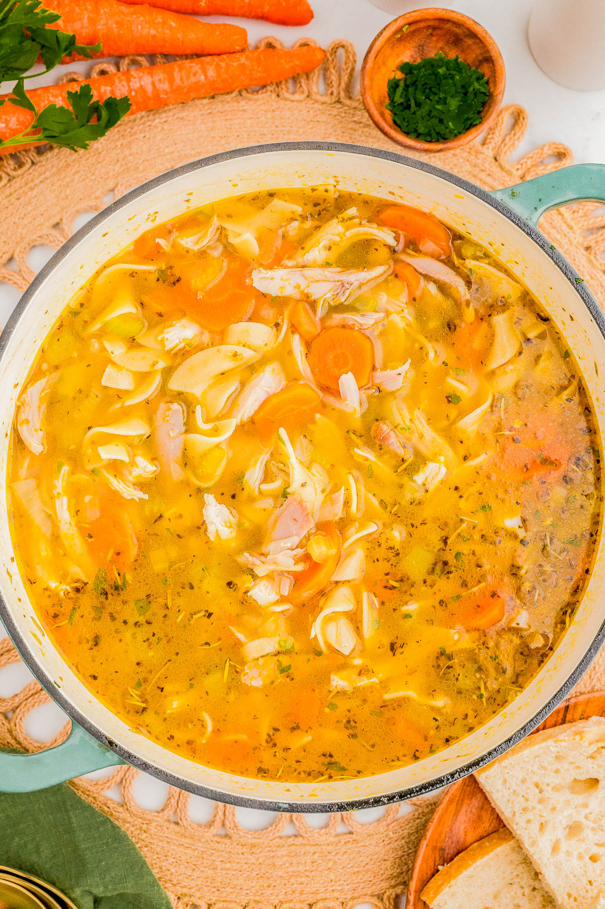 A pot of chicken noodle soup with sliced carrots, chicken pieces, and herbs, surrounded by parsley, carrots, and bread.