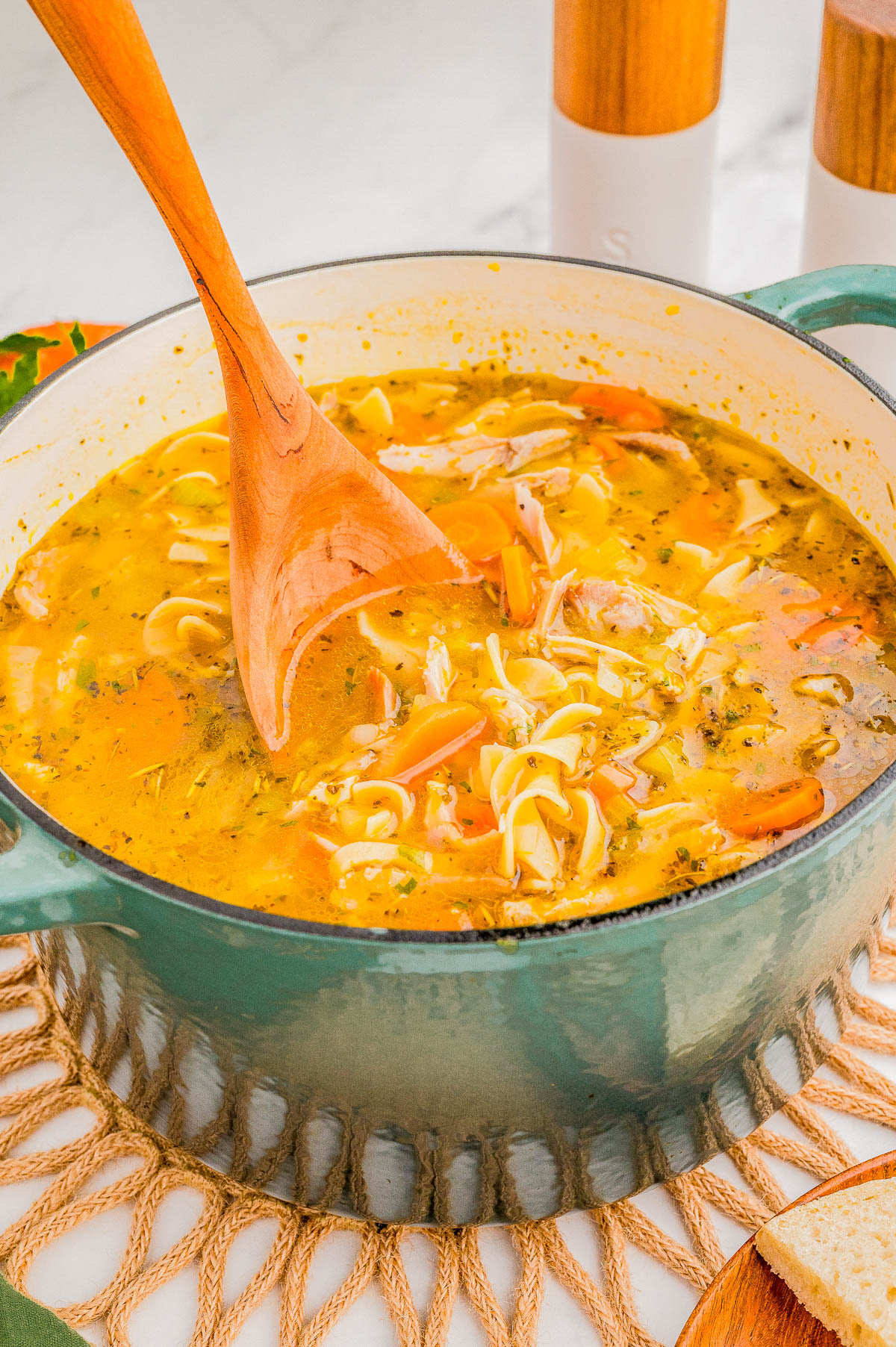 A pot of chicken noodle soup with carrots and herbs being stirred with a wooden spoon.