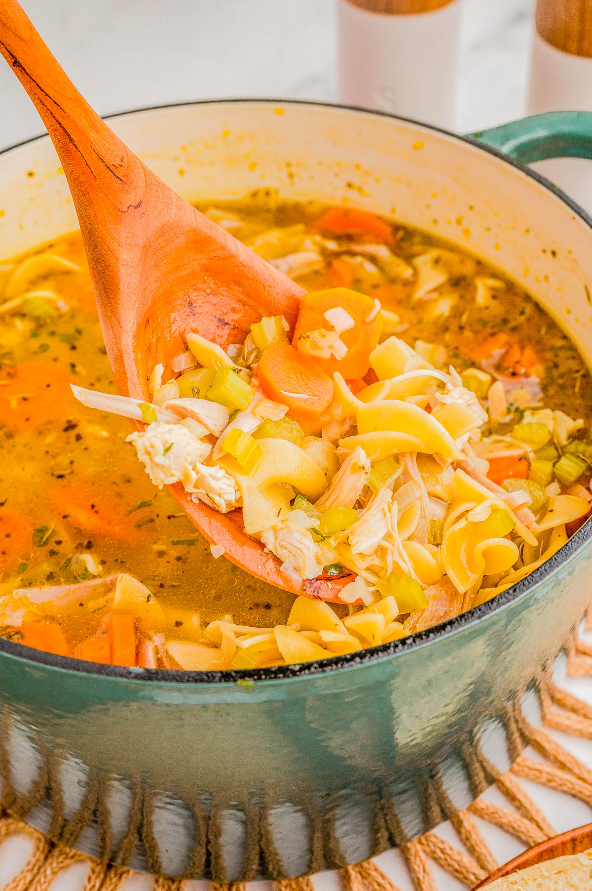 A wooden spoon lifts chicken noodle soup with vegetables from a green pot.