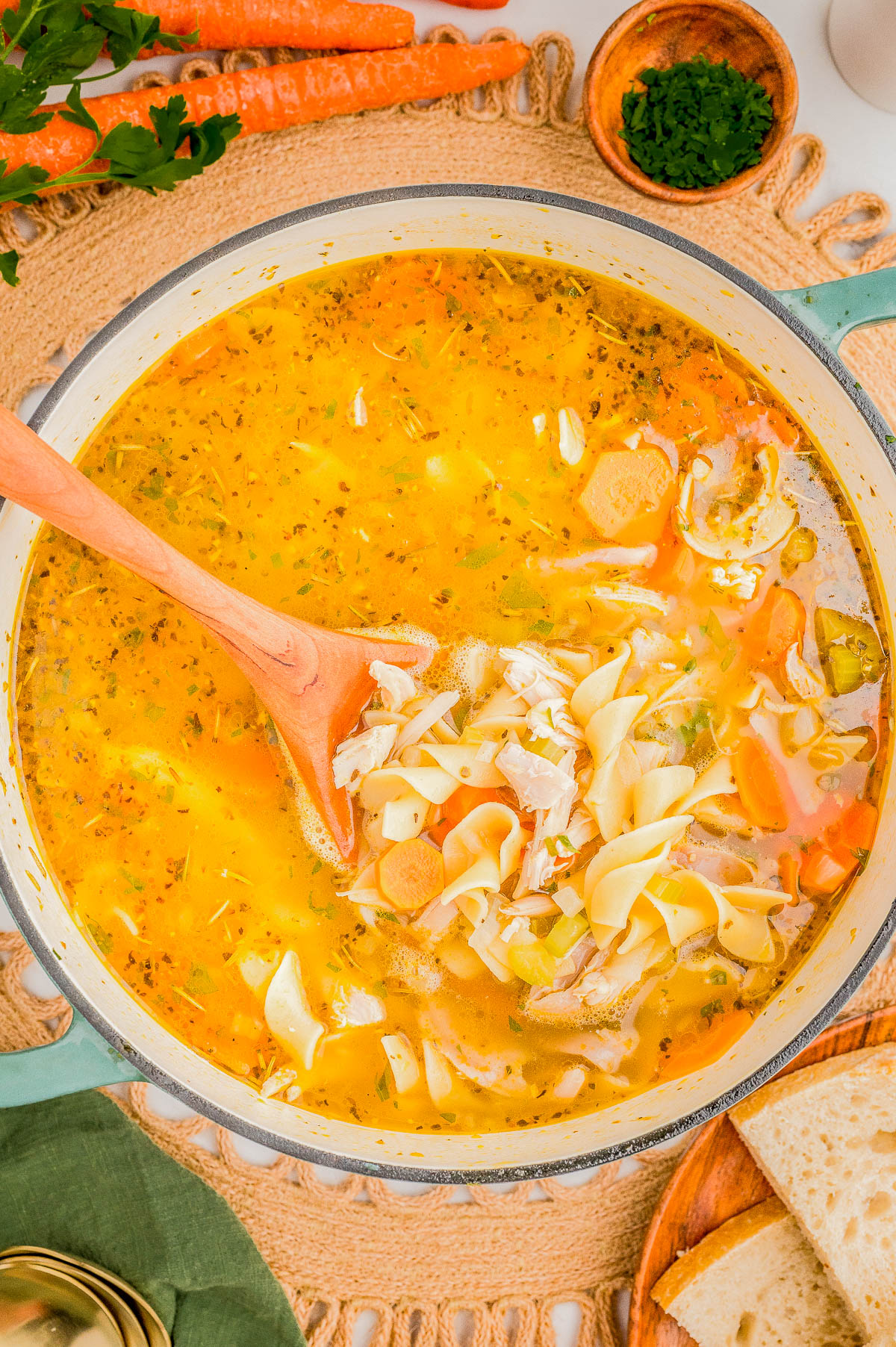 A pot of homemade chicken noodle soup with a wooden spoon, surrounded by carrots, parsley, and slices of bread on a woven mat.