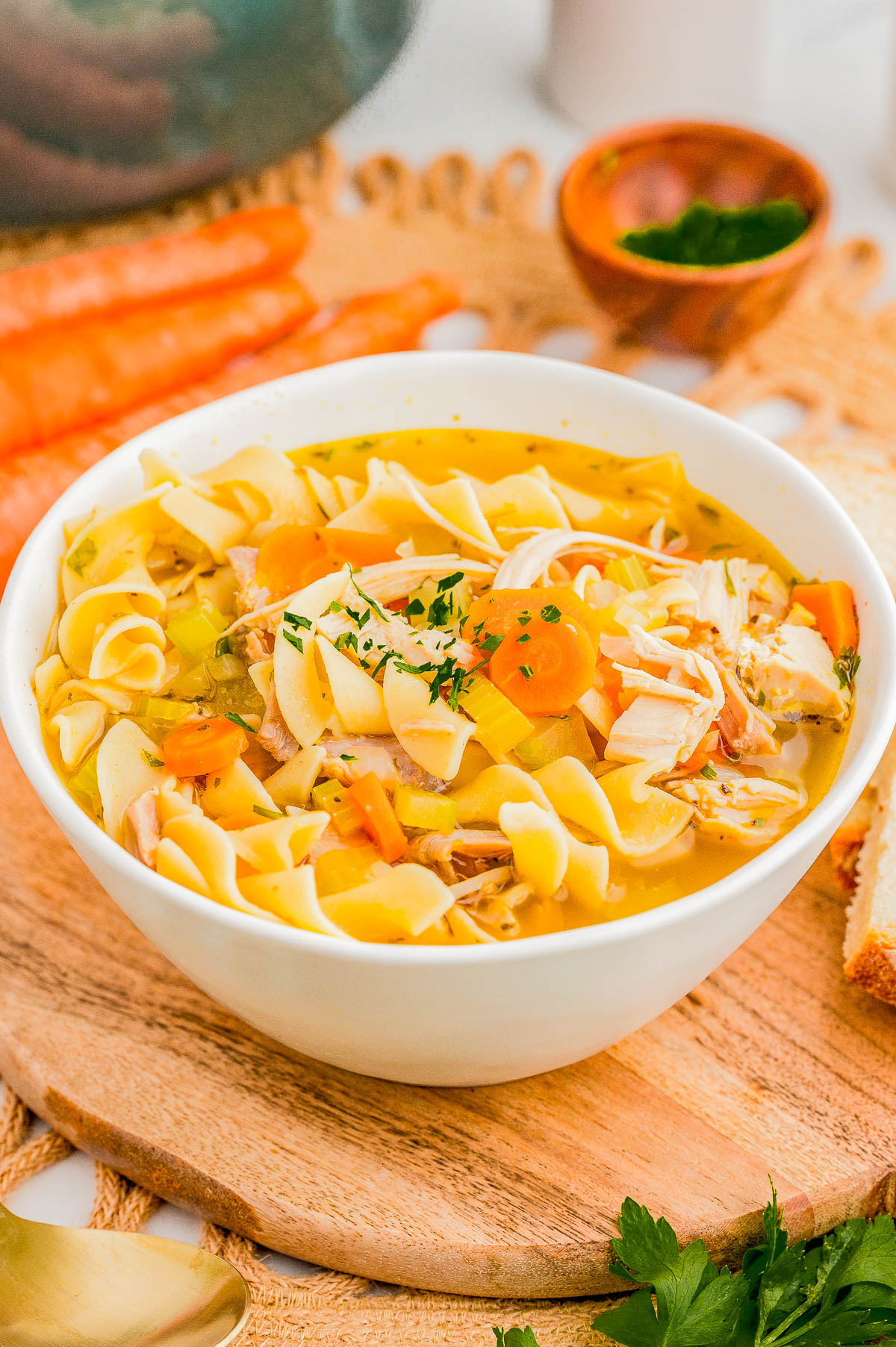 A bowl of chicken noodle soup with carrots and herbs on a wooden board, surrounded by fresh carrots and greens.