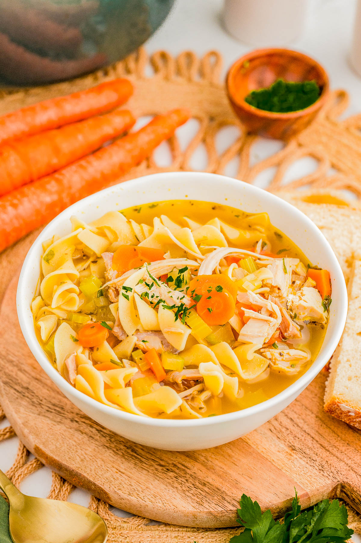 A bowl of chicken noodle soup with carrots and herbs, placed on a wooden board with slices of bread, fresh carrots, and parsley in the background.