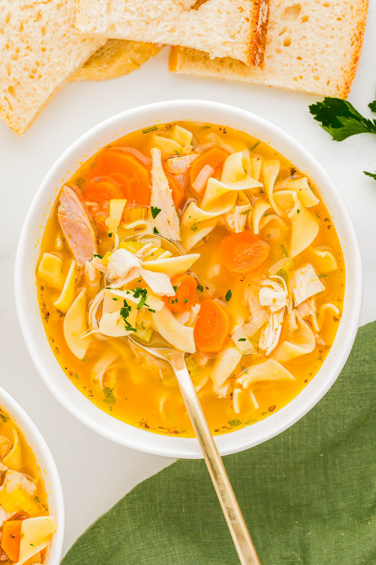 A bowl of chicken noodle soup with carrots, noodles, and parsley is placed on a table. Slices of bread are in the background.