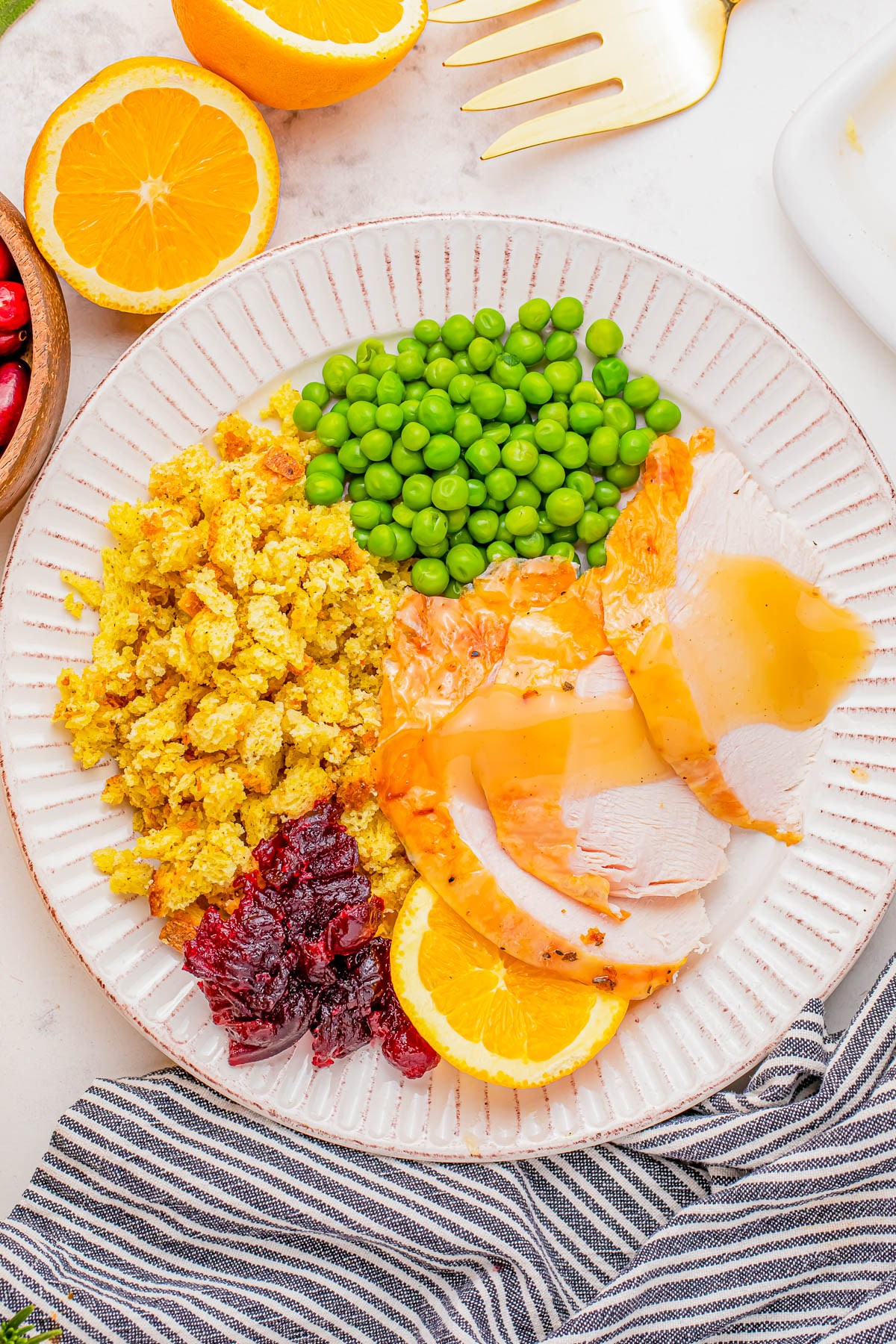 A plate with sliced turkey, peas, stuffing, cranberry sauce, and orange slices. A fork, striped napkin, and orange garnish are also visible.