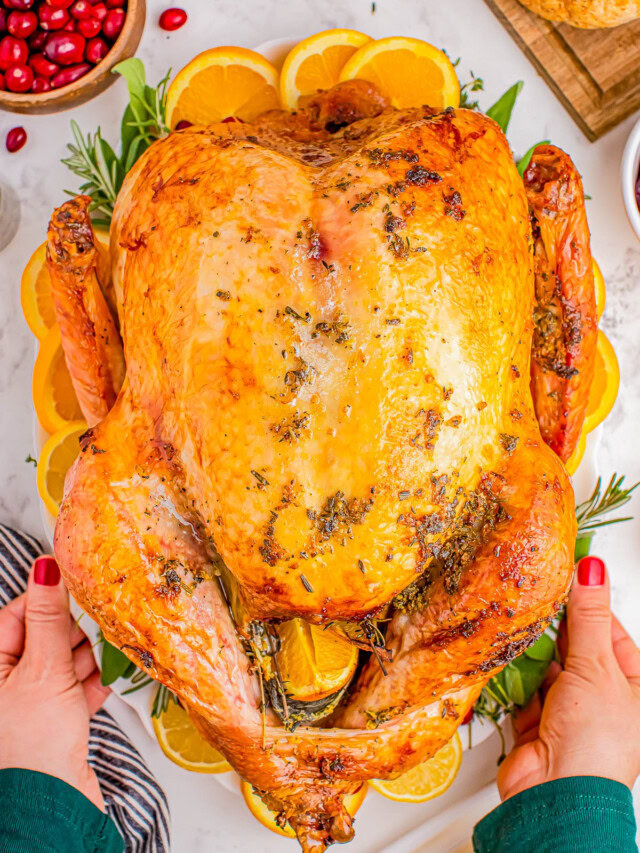 A whole roasted turkey garnished with herbs and lemon slices, held by a person with red nail polish. Cranberries and peas are visible in the background.