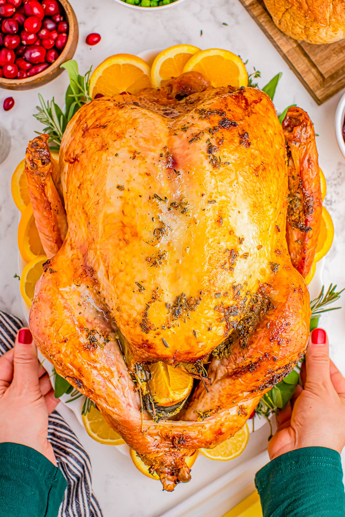 A whole roasted turkey garnished with herbs and lemon slices, held by a person with red nail polish. Cranberries and peas are visible in the background.