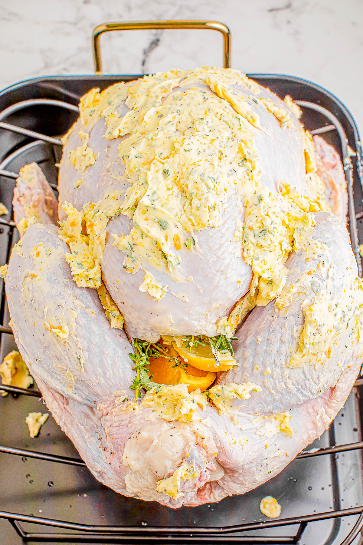 Uncooked turkey on a roasting rack, covered with herb butter and stuffed with herbs and lemon slices, ready for roasting.