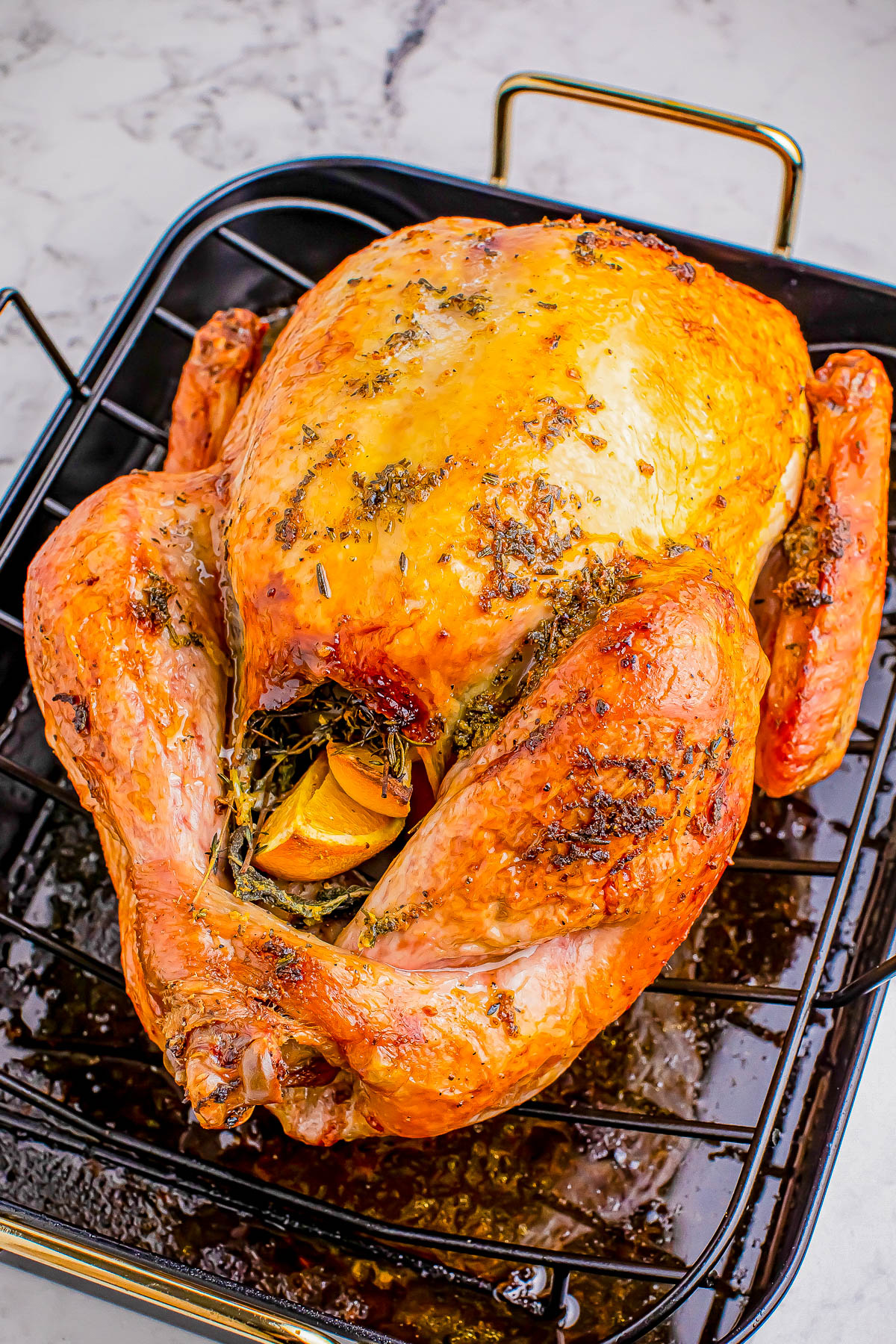 Roasted turkey with herbs and lemon slices inside, placed on a metal rack over a roasting pan.