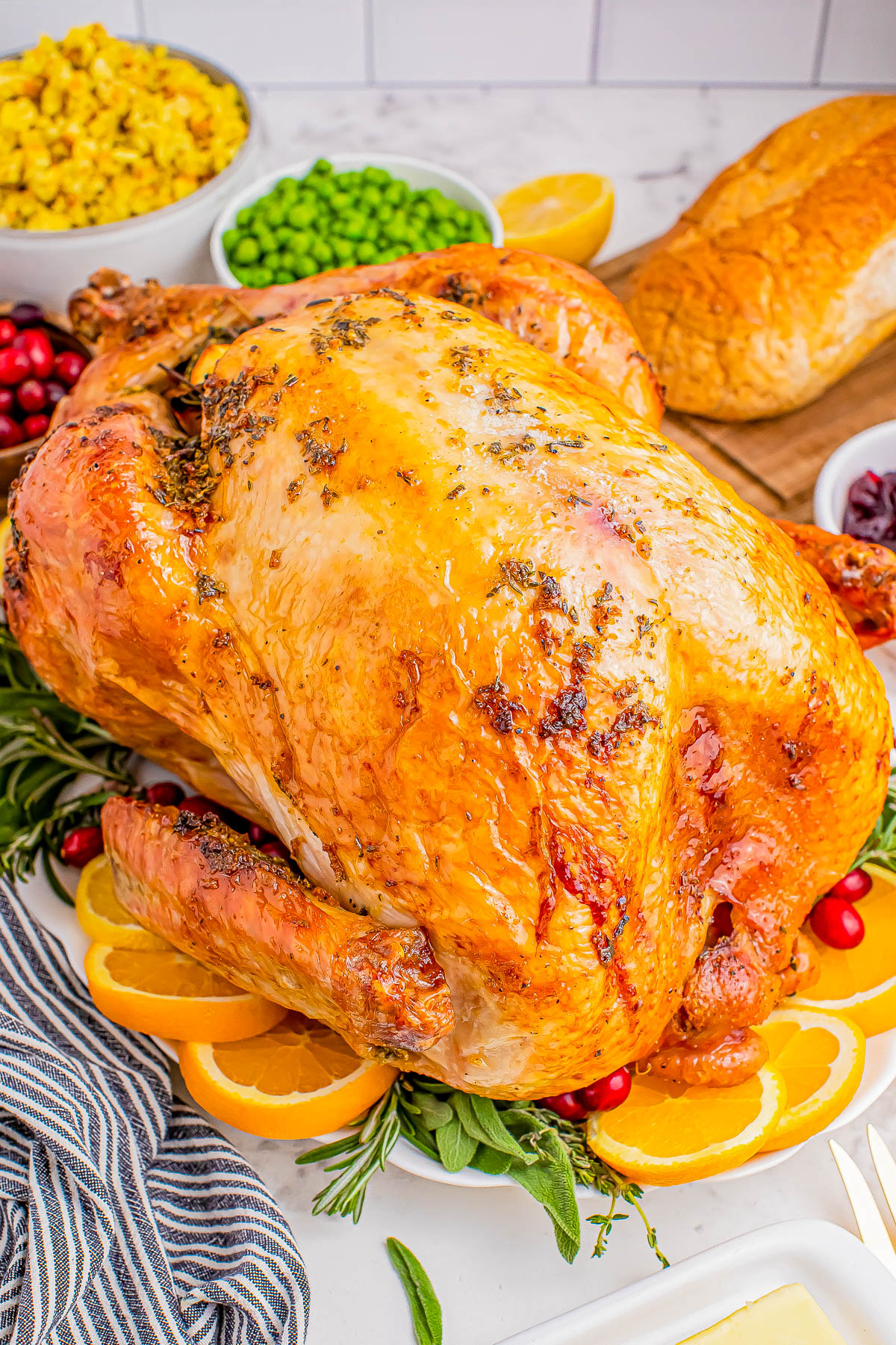 A golden-brown roasted turkey garnished with orange slices and herbs, surrounded by bowls of peas, corn, cranberries, and a loaf of bread on a white surface.