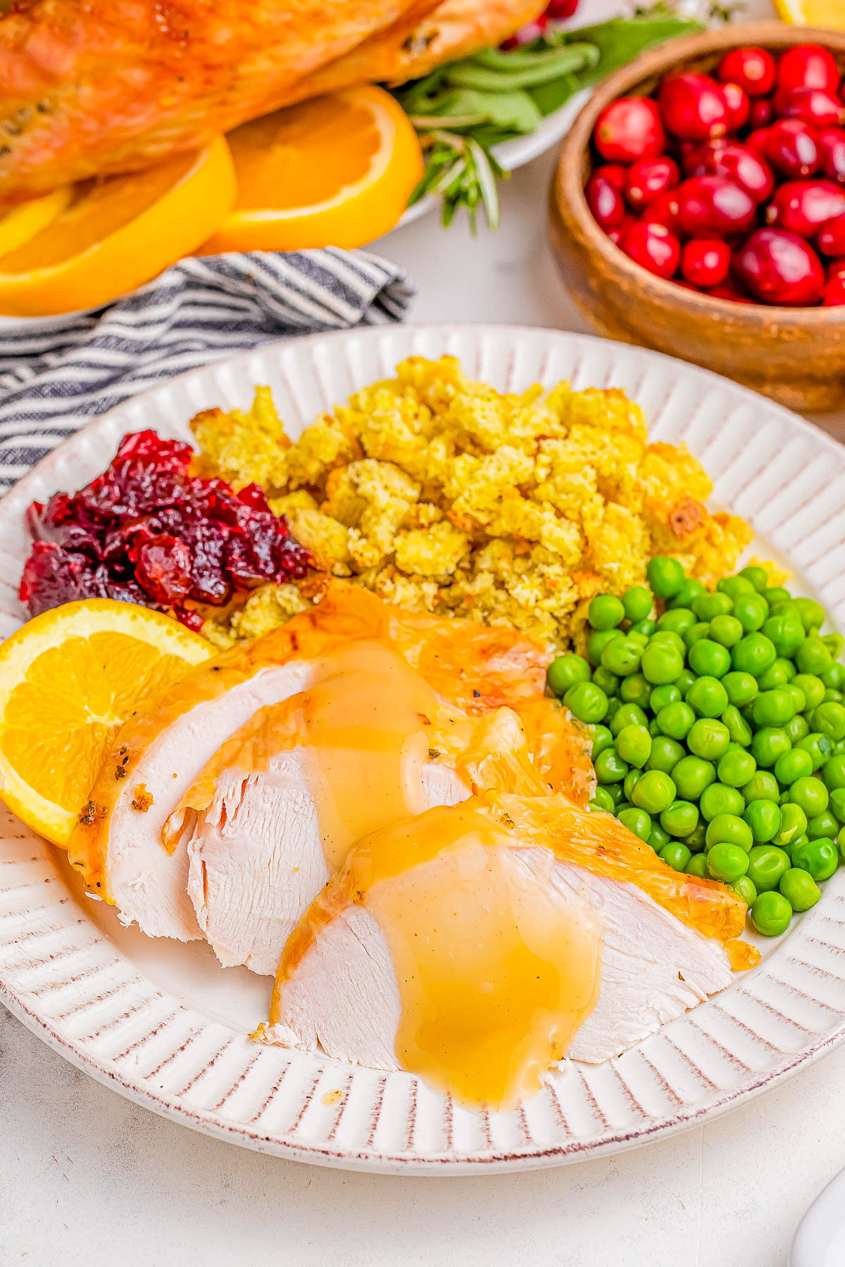 Plate with sliced turkey and gravy, peas, stuffing, cranberry sauce, and an orange slice. Bowl of cranberries in the background.