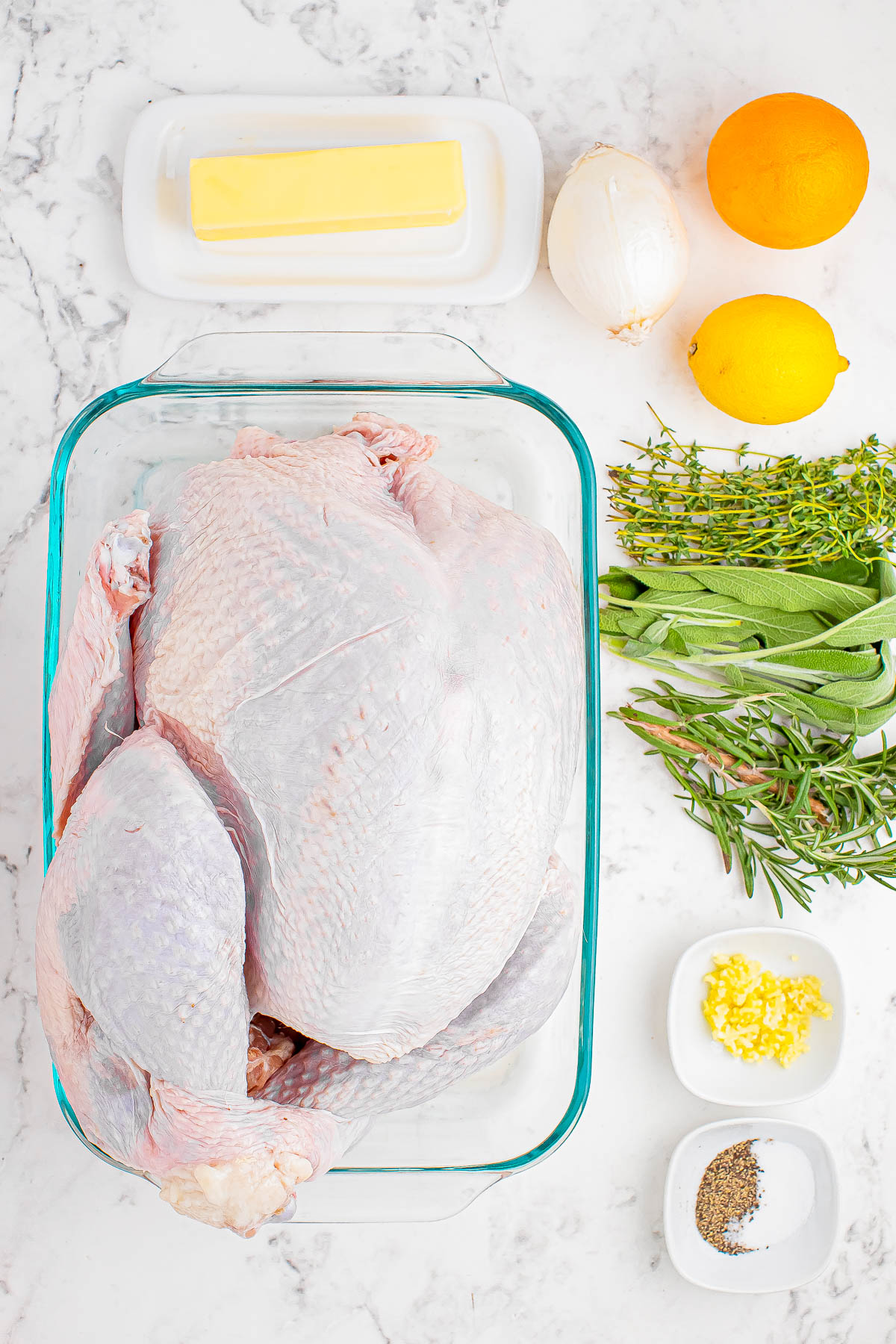 A raw turkey in a glass dish surrounded by butter, oranges, lemon, an onion, fresh herbs, minced garlic, and spices on a marble countertop.