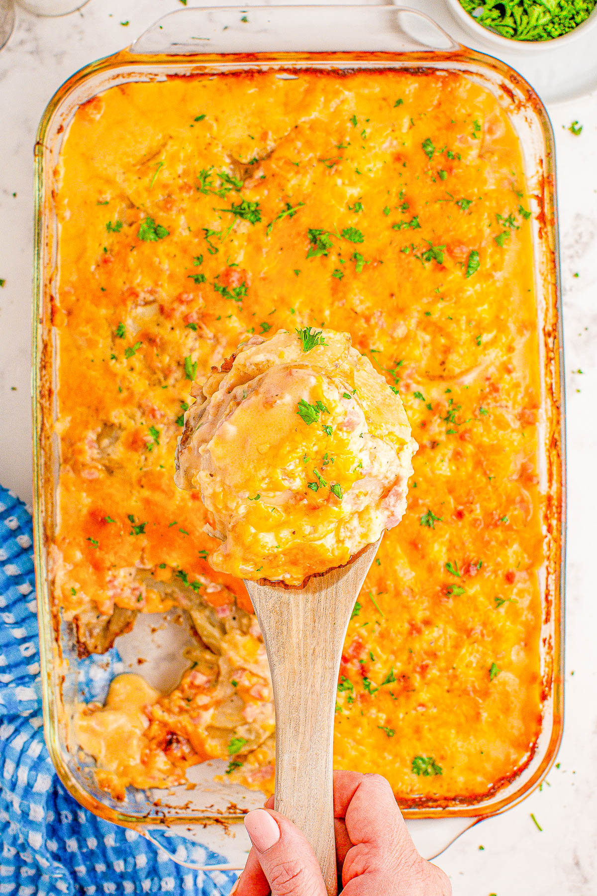 A hand holding a wooden spoon serves a portion of baked cheesy casserole garnished with parsley, with a blue and white cloth nearby.