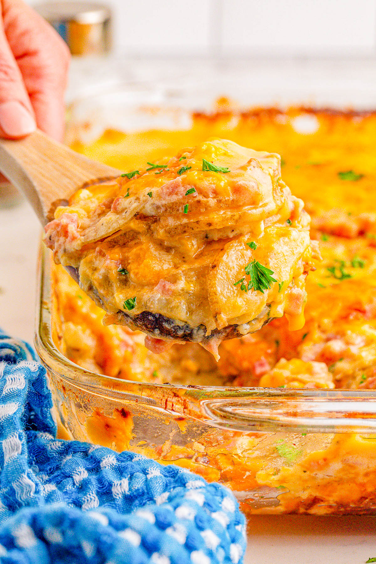 A hand holds a wooden spatula lifting a portion of cheesy casserole with vegetables from a glass baking dish. A blue and white towel is nearby.