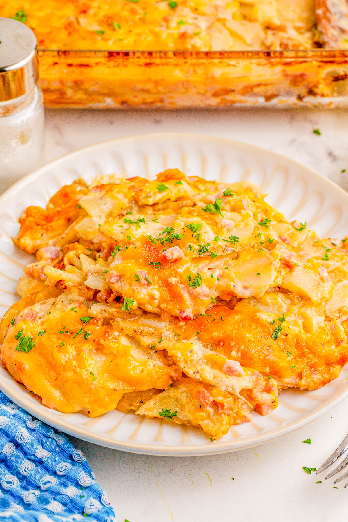 A plate of cheesy scalloped potatoes topped with parsley, with a casserole dish visible in the background.