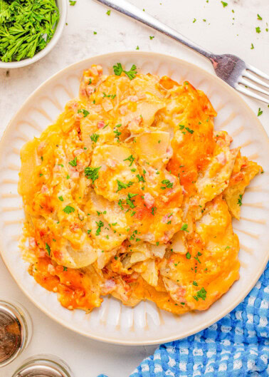 A plate of cheesy scalloped potatoes garnished with parsley, next to a fork, a blue cloth, and a small dish of fresh herbs.