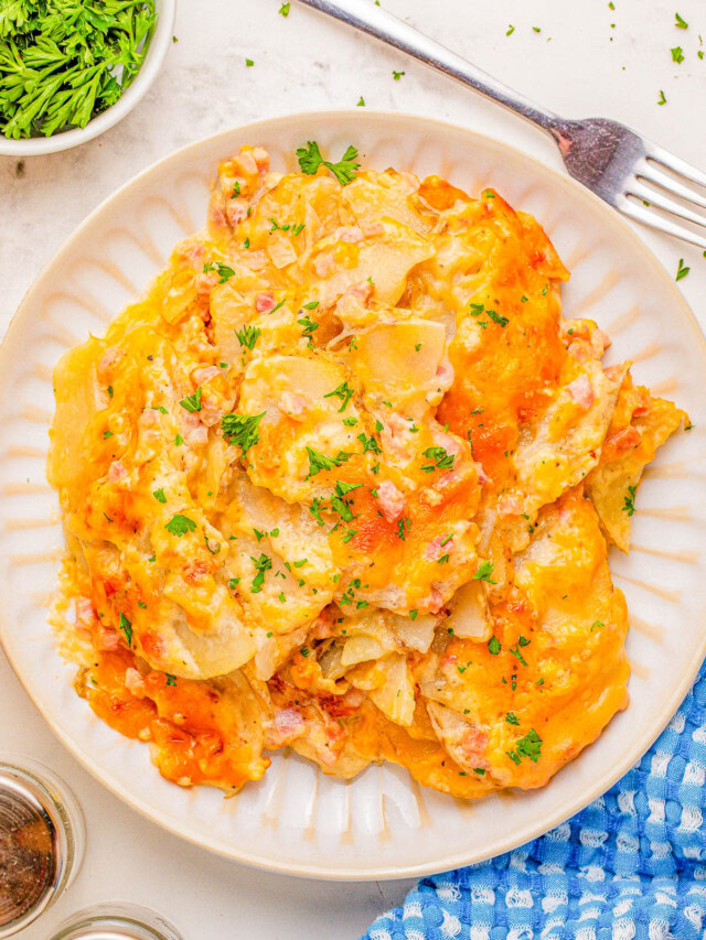 A plate of cheesy scalloped potatoes garnished with parsley, next to a fork, a blue cloth, and a small dish of fresh herbs.