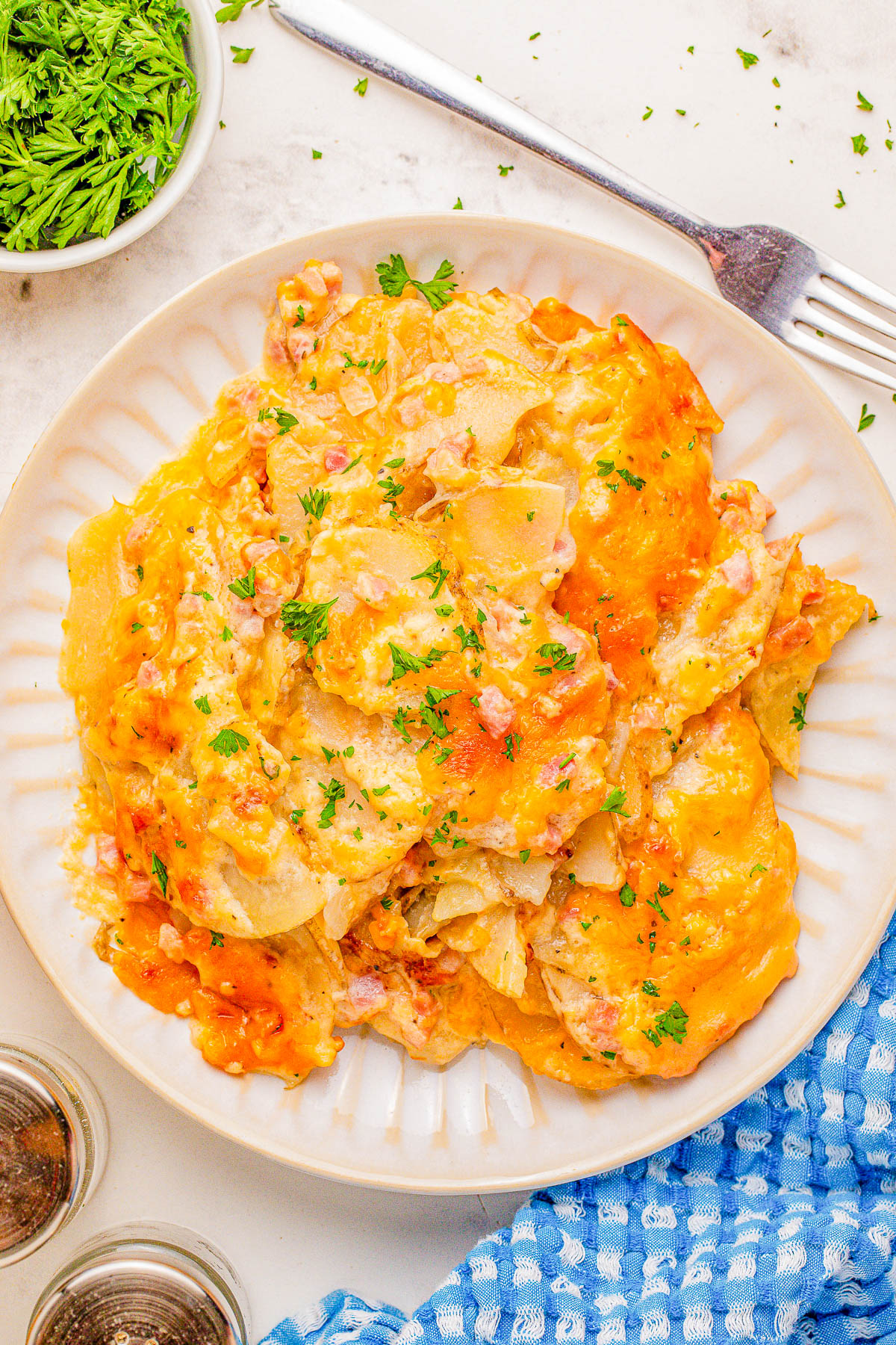 A plate of cheesy scalloped potatoes garnished with parsley, next to a fork, a blue cloth, and a small dish of fresh herbs.