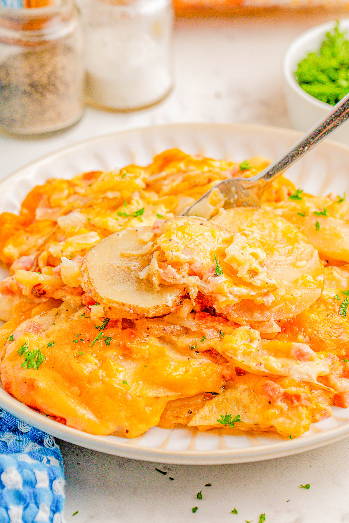 A plate of cheesy scalloped potatoes with a fork on top, garnished with parsley. Salt and pepper shakers and a small bowl of herbs are in the background.