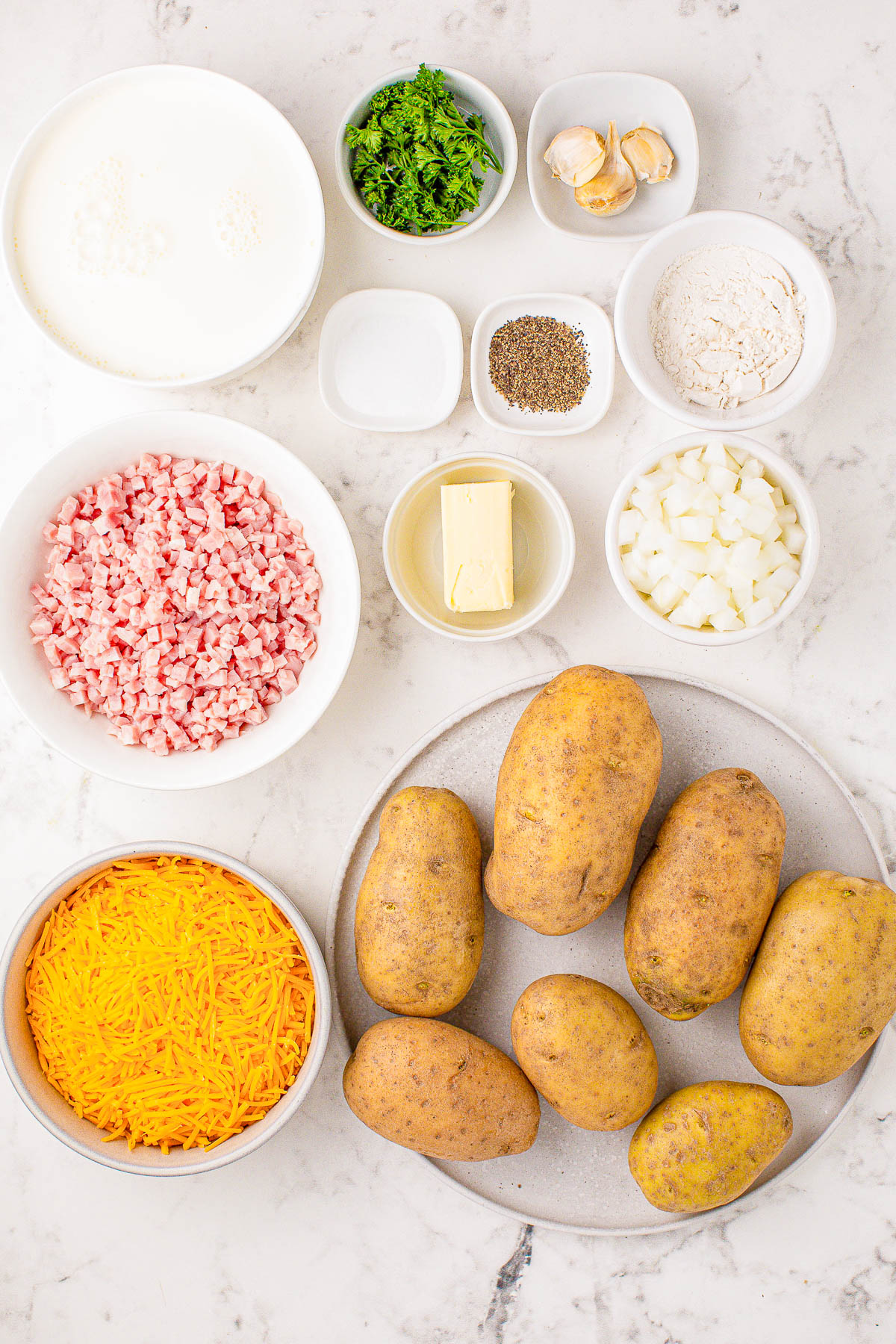 Ingredients on a countertop include potatoes, shredded cheese, diced ham, chopped onions, garlic, butter, flour, milk, cream, parsley, black pepper, and salt.