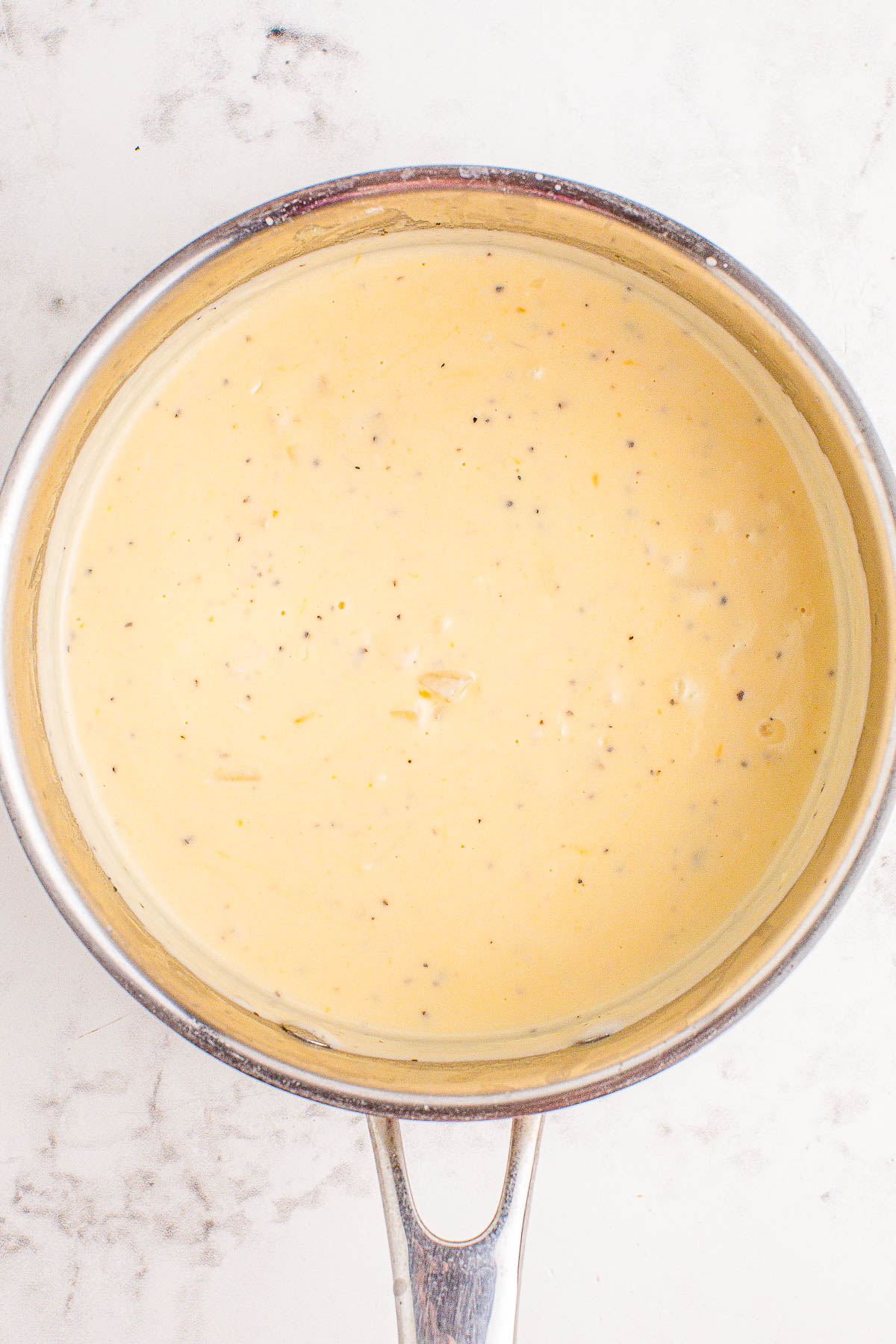 A saucepan filled with creamy, light yellow cheese sauce with visible black pepper flakes, placed on a light-colored surface.