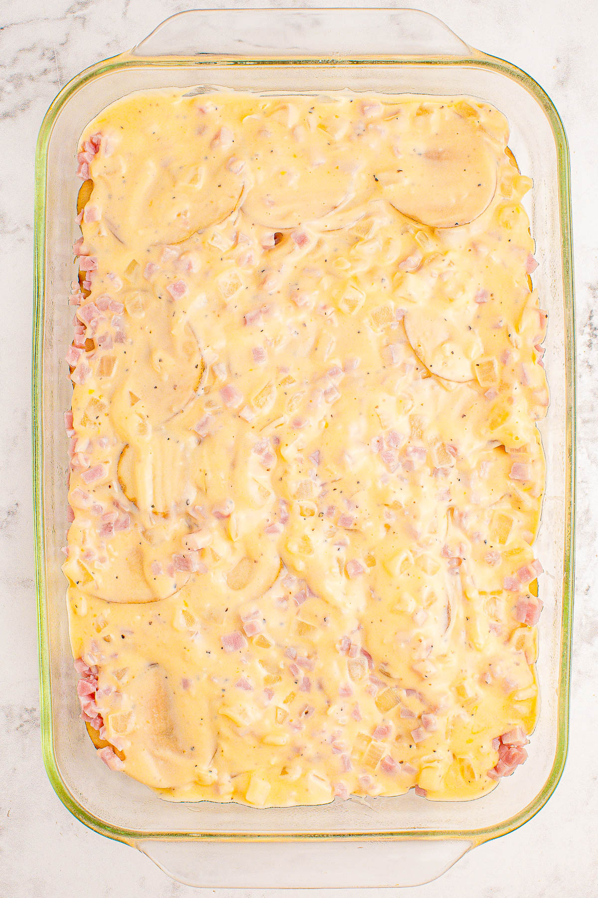 A glass baking dish filled with a creamy, layered casserole, featuring thinly sliced potatoes, diced ham, and melted cheese.