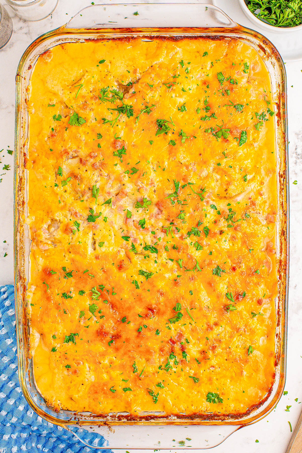 A baked casserole dish topped with melted cheese and sprinkled with chopped parsley, placed on a marble countertop next to a blue checkered cloth.