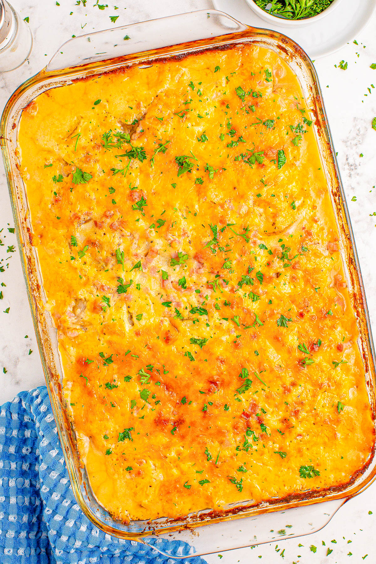 A baked casserole topped with melted cheese and sprinkled with chopped parsley, placed on a countertop next to a blue cloth.