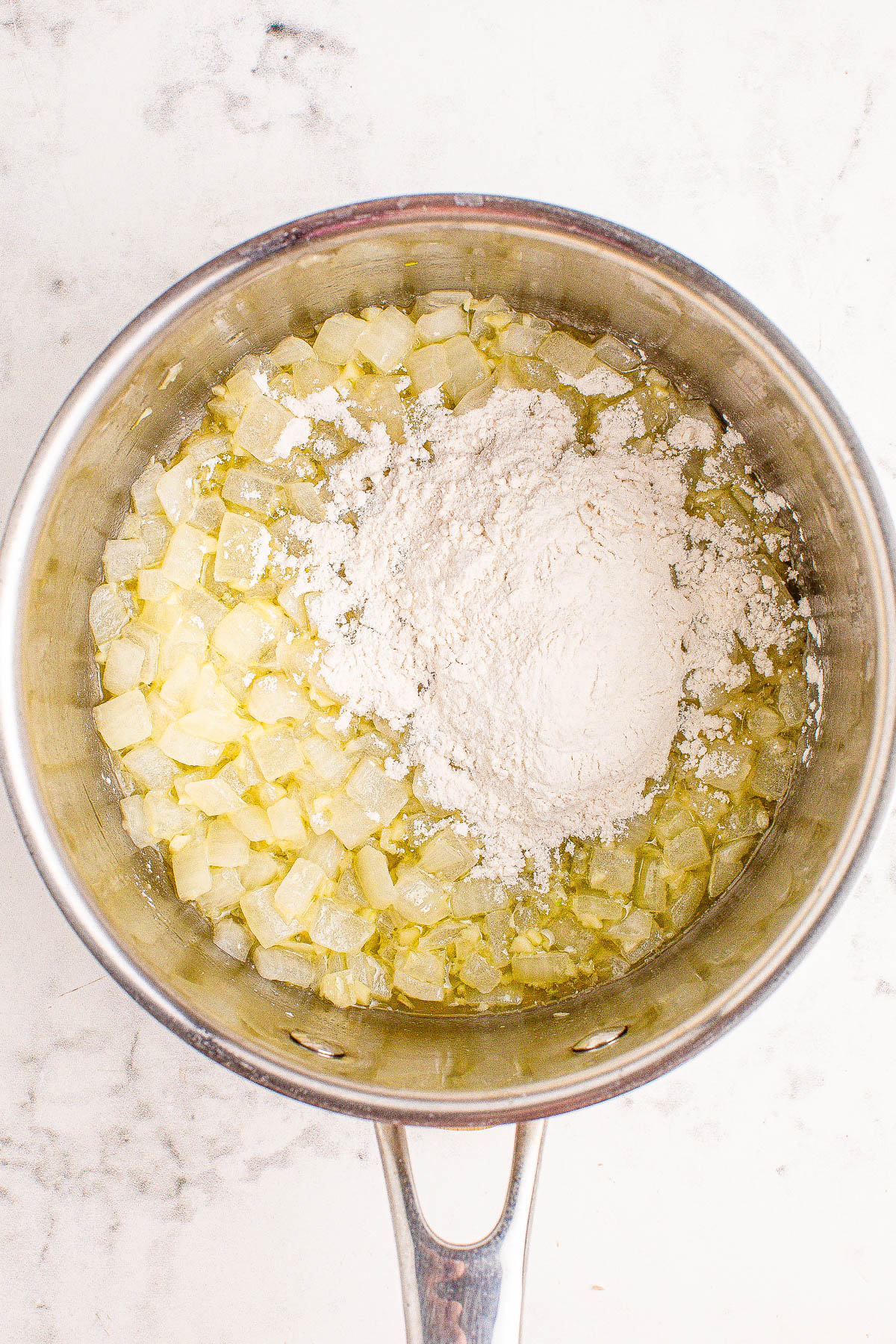 Chopped onions and flour in a stainless steel pot on a marble countertop.