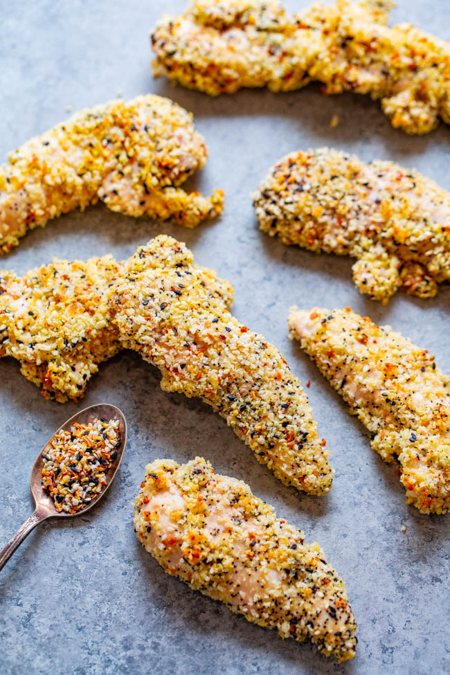 Breaded chicken tenders arranged on a gray surface with a spoonful of seasoning mixture beside them.