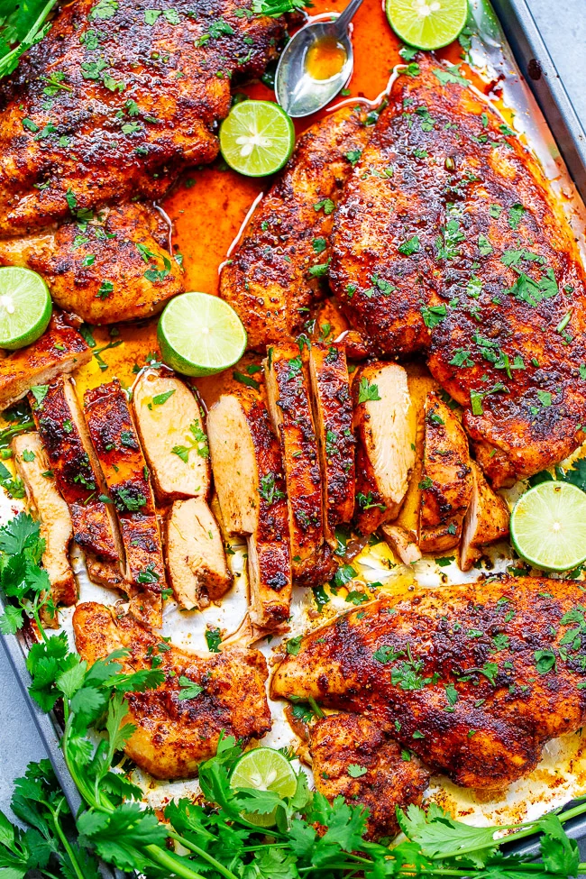 A baking sheet with seasoned, sliced roast chicken topped with herbs, slices of lime, and a spoon. Surrounding it are fresh herbs and lime halves.