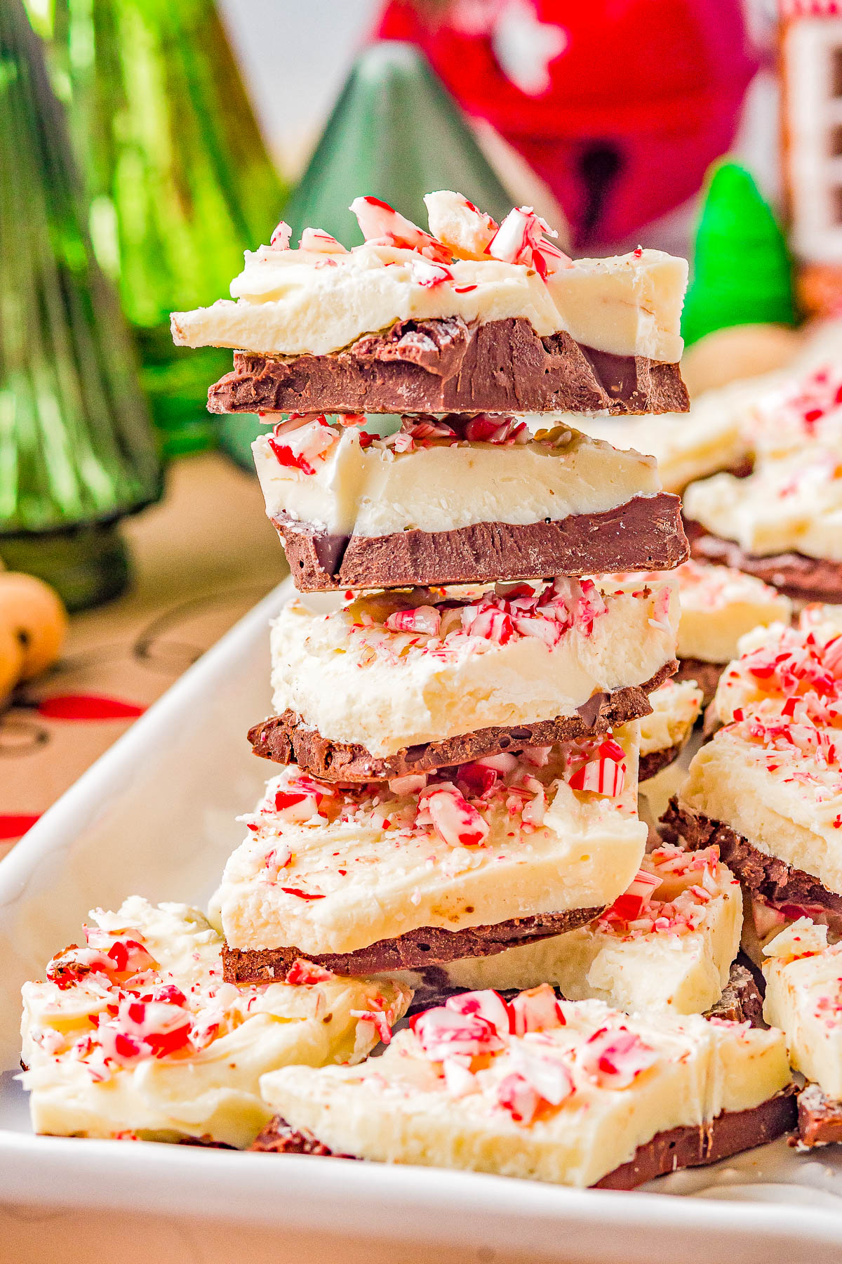 A stack of peppermint bark pieces with layers of chocolate and white chocolate, topped with crushed candy canes, on a white plate. Holiday decorations are visible in the background.