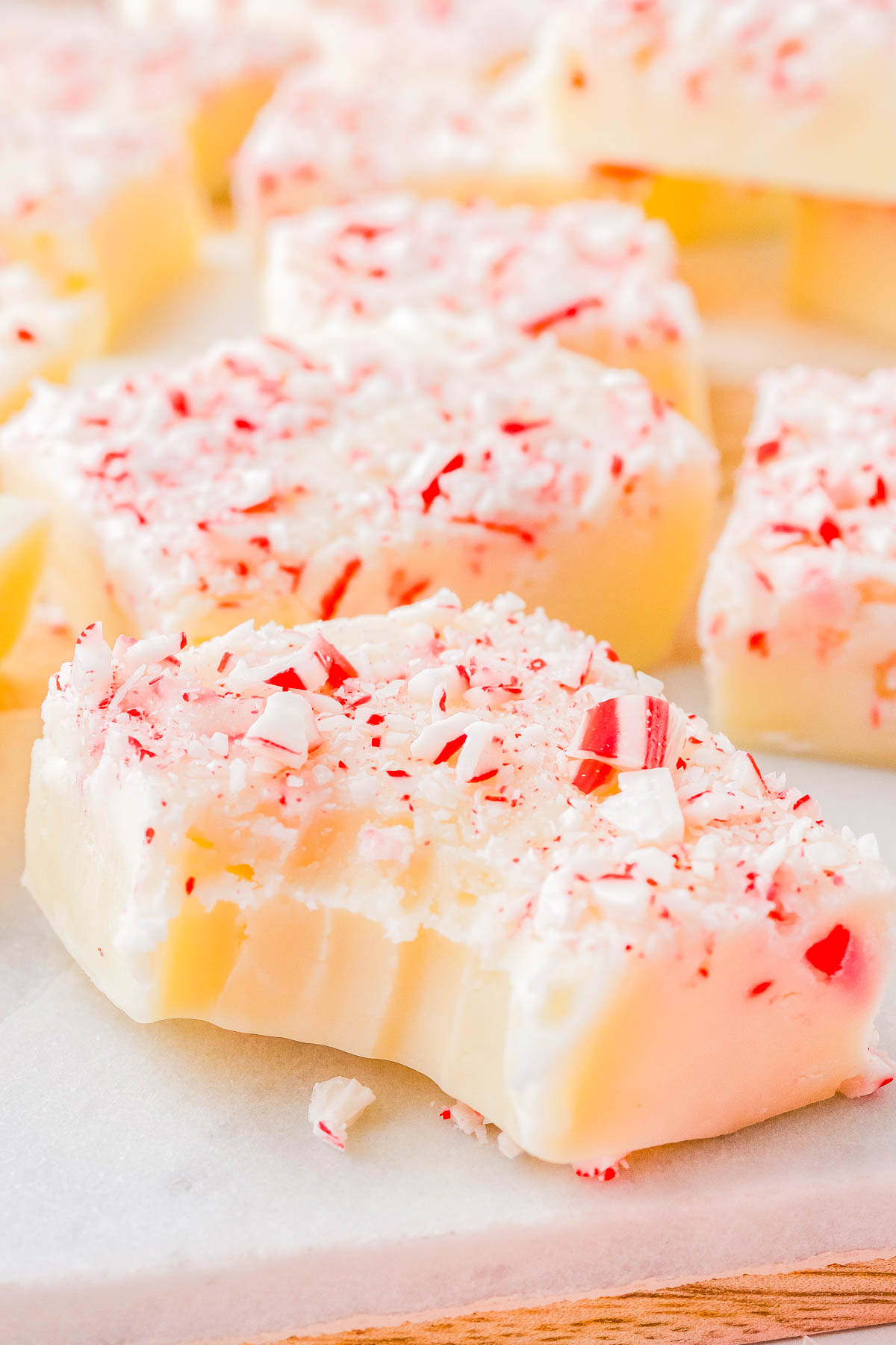 Close-up of white fudge pieces topped with crushed peppermint candy on a white surface. One piece shows a bite taken out.