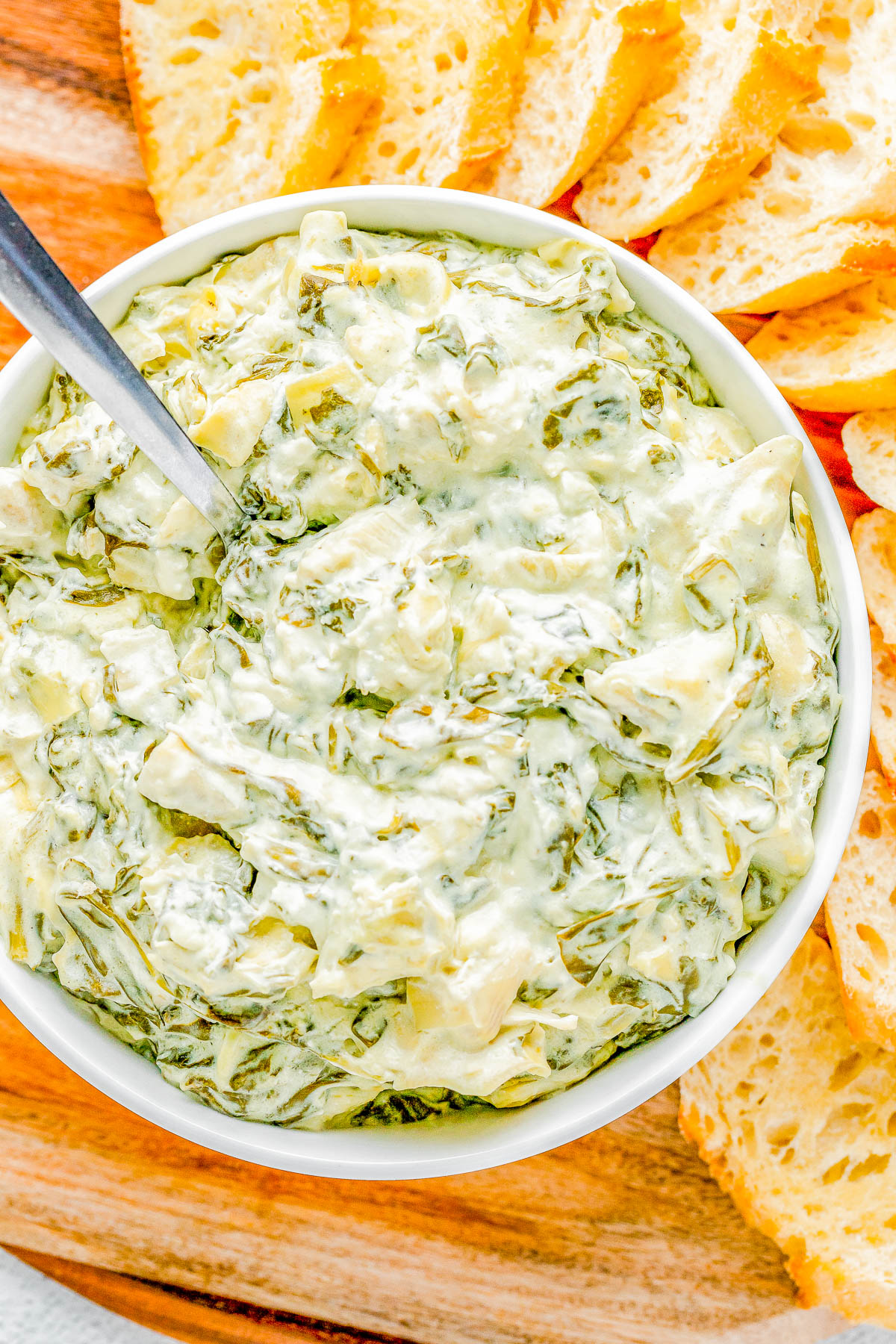 A bowl of creamy spinach and artichoke dip with a spoon, surrounded by slices of bread on a wooden board.