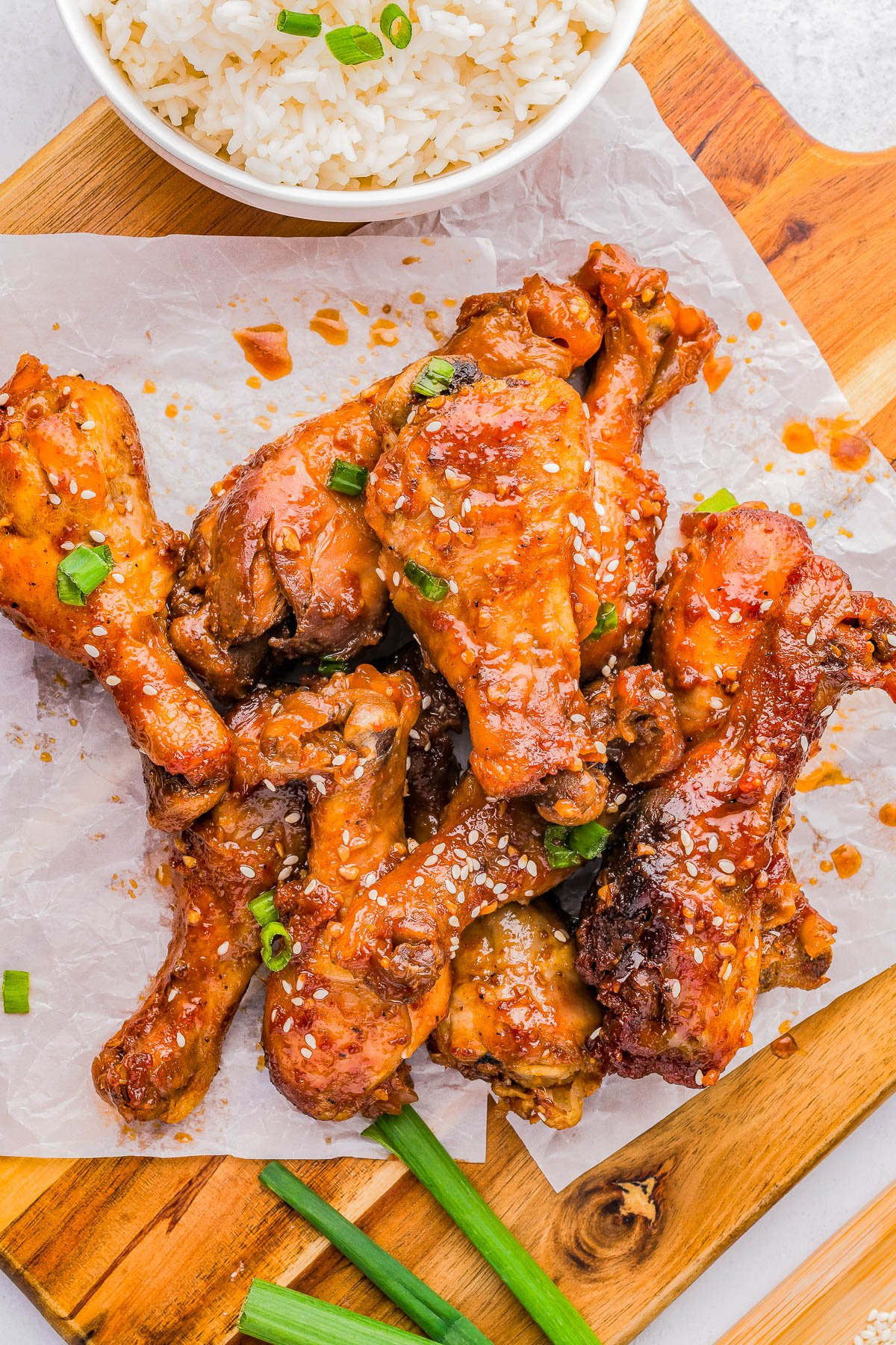 Chicken drumsticks glazed with sauce, garnished with sesame seeds and green onions, served on a wooden board with a bowl of rice.