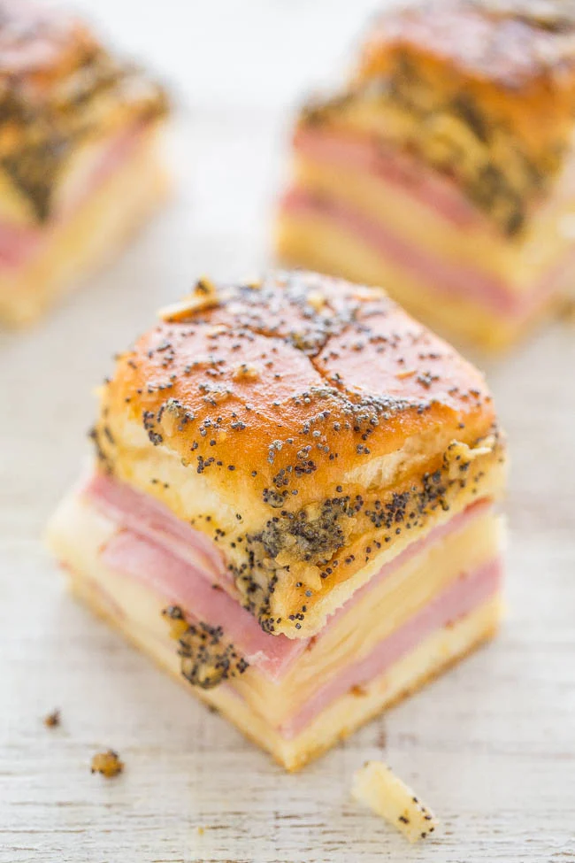 Close-up of a small layered sandwich with poppy seed topping, containing ham and cheese, on a light wooden surface.