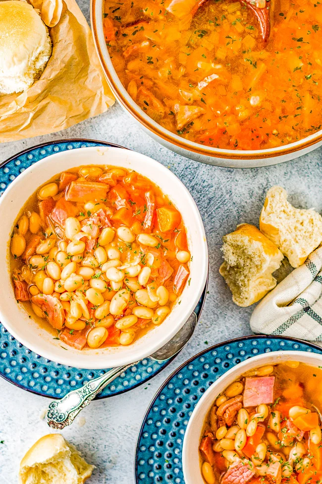 Bowls of bean and ham soup with a spoon on patterned plates, bread pieces, and a pot with more soup.