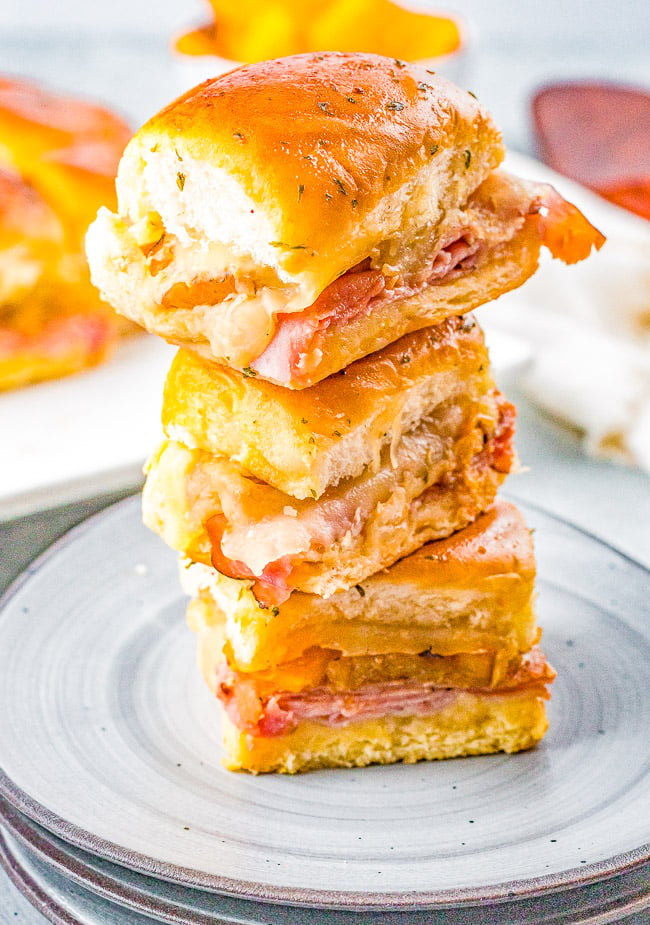 A stack of three cheesy ham and tomato sliders on a plate, topped with a golden bun.