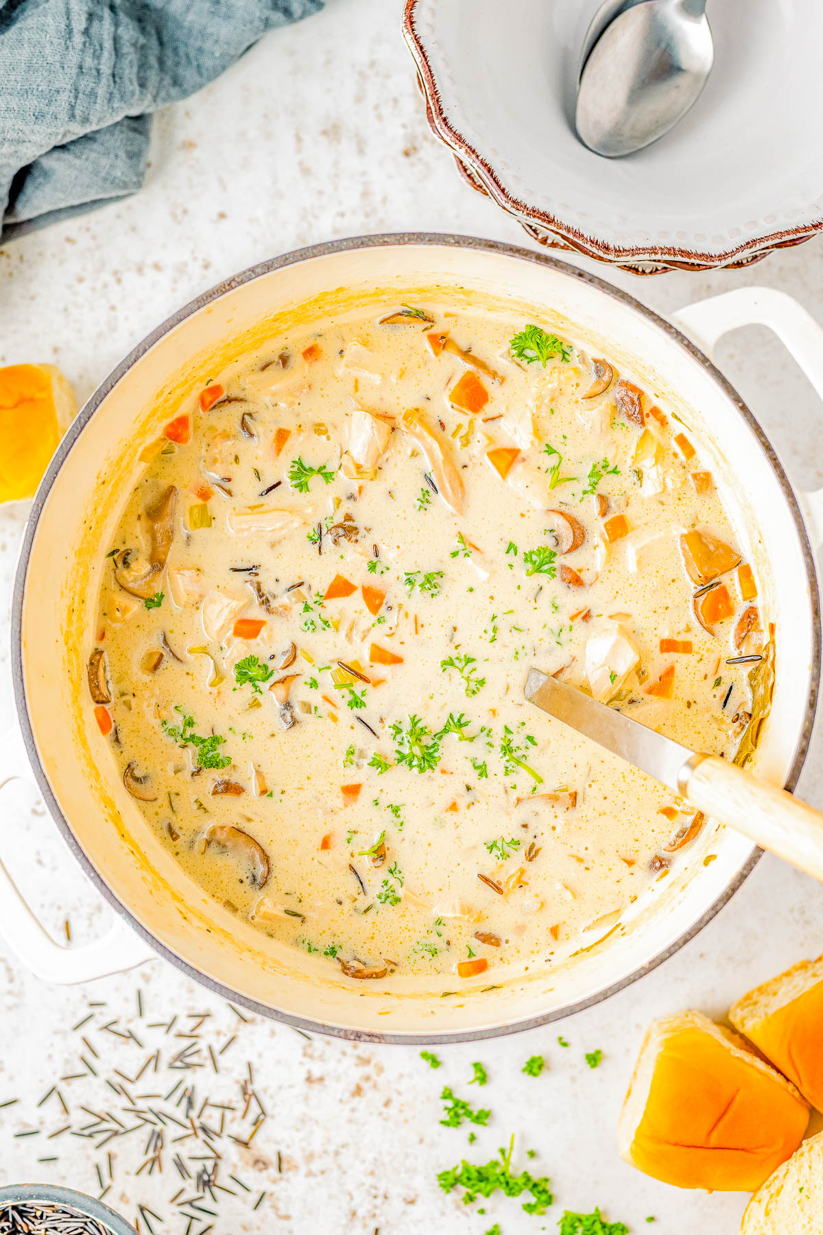 A pot of creamy soup containing diced vegetables and herbs, with a ladle resting inside. Nearby are sliced pumpkins and a bowl with spoons.