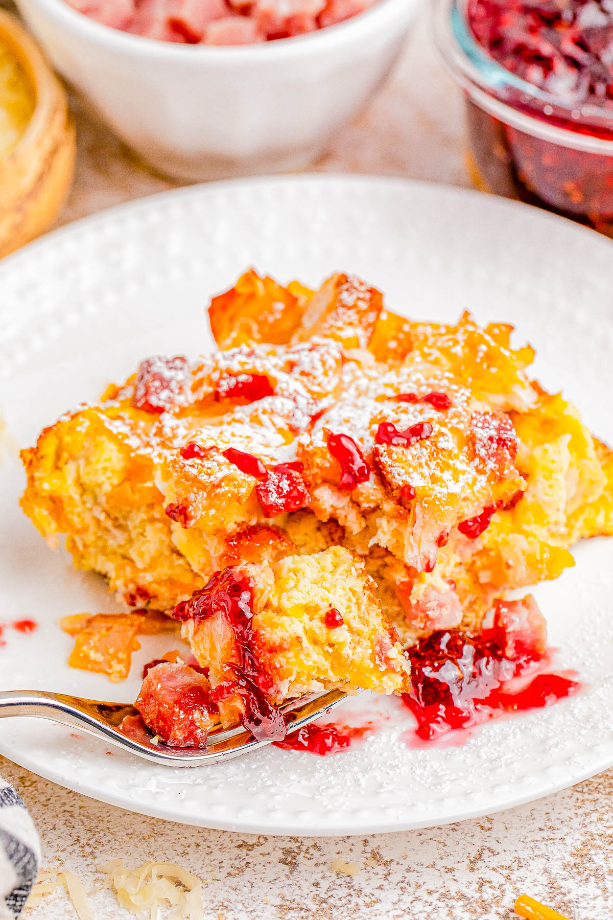 A slice of bread pudding on a plate, topped with powdered sugar and fruit sauce, with a fork resting on the side.