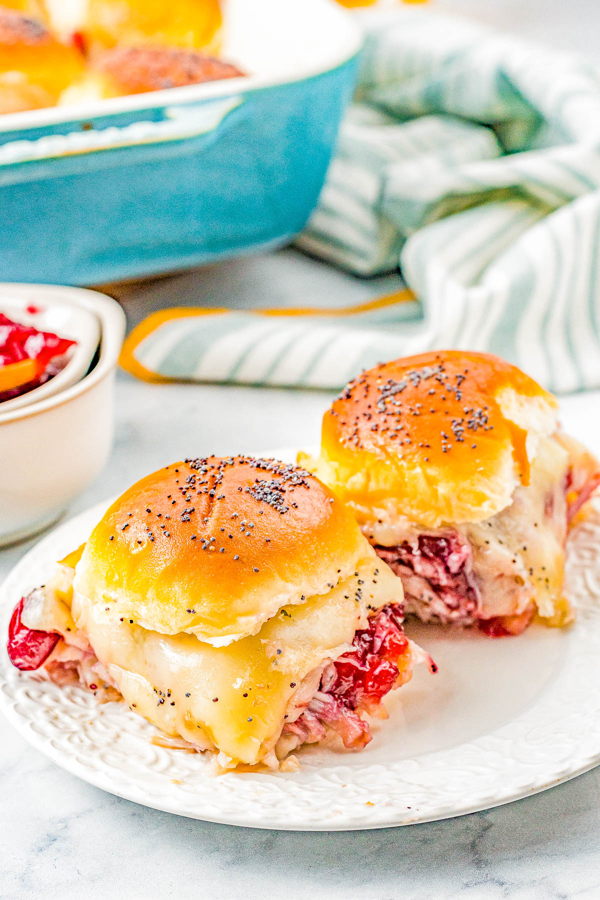 Two small slider sandwiches with melted cheese and poppy seeds on top, filled with turkey and cranberry sauce, on a white plate. A baking dish and striped cloth are in the background.