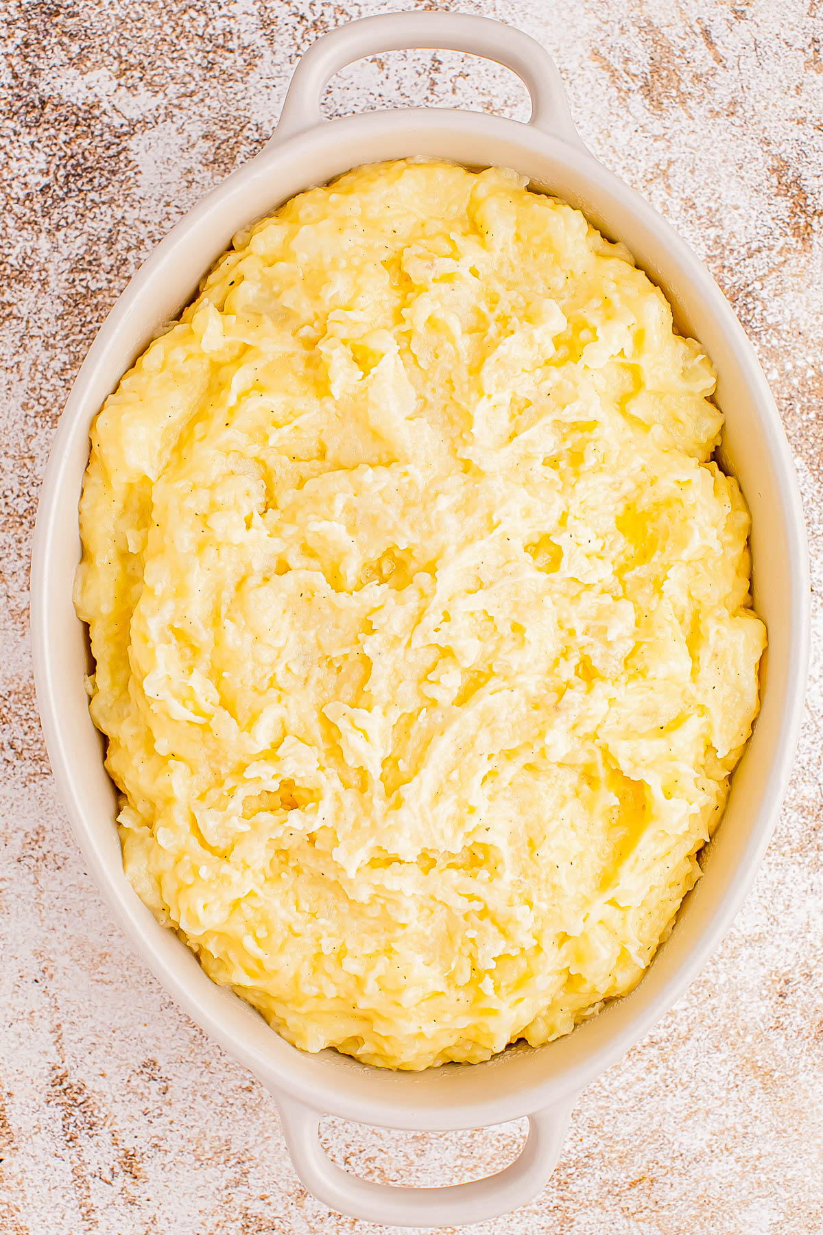 A white oval dish filled with creamy mashed potatoes on a textured surface.