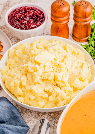 A bowl of mashed potatoes with butter on top, surrounded by bowls of cranberry sauce and gravy, with salt and pepper shakers in the background.