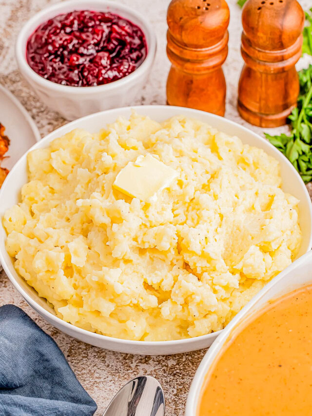 A bowl of mashed potatoes with butter on top, surrounded by bowls of cranberry sauce and gravy, with salt and pepper shakers in the background.