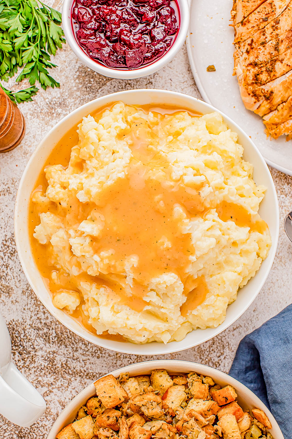 A bowl of mashed potatoes topped with gravy, surrounded by cranberry sauce, sliced turkey, and stuffing.